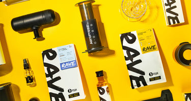 Coffee brewing items, coffee bags labeled “RAVE,” an AeroPress, grinder, cups, and small bottles are arranged on a bright yellow background. The layout appears organized and vibrant.