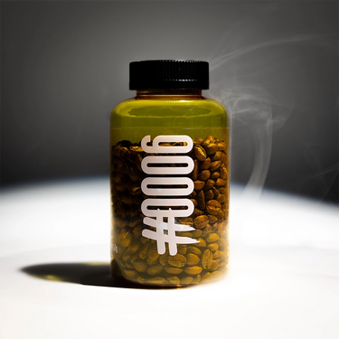 A clear cylindrical bottle with a black cap, filled with roasted coffee beans, standing on a white surface against a dark gray background with a smoky effect. Large white text on the bottle reads “0006” in a stylized vertical font.