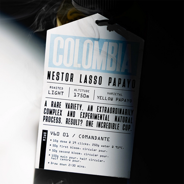 A close-up of a white coffee bag label reading 'COLOMBIA NESTOR LASSO PAPAYO' with roast, altitude, variety, brewing instructions, and a small coffee bean illustration, photographed against a dark background with a blurred coffee bean bag visible behind it.