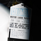A close-up of a white coffee bag label reading 'COLOMBIA NESTOR LASSO PAPAYO' with roast, altitude, variety, brewing instructions, and a small coffee bean illustration, photographed against a dark background with a blurred coffee bean bag visible behind it.