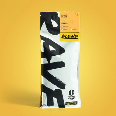 A bag of Rave Coffee’s Fudge Blend Nº 5 sits on a yellow background. The white packaging with bold black text shows tasting notes and a “1% for the Planet” label—ideal for your next flat white or cappuccino.