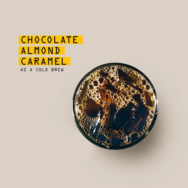 A glass of iced Cold Brew Ground Coffee Nº. 9 viewed from above, with visible ice cubes and bubbles. Text beside the glass reads: Chocolate Almond Caramel as a cold brew coffee on a light beige background.