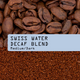 A close-up of whole, decaffeinated coffee beans above ground coffee with a label: Swiss Water Decaf Blend Nº 11, Medium/Dark—crafted using the natural Swiss water process decaf method.
