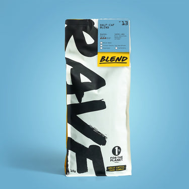 A bag of Half Caf Blend Nº 13 medium roast coffee stands upright on a blue background. The white packaging features bold black RAVE text, a blue label at the top, and a yellow accent highlighting 1% for the planet.