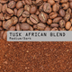 A close-up of whole Rwandan coffee beans above and ground coffee below, divided by a gray label reading Tusk African Blend Nº 24 in bold black text. This medium-dark roast supports conservation efforts.