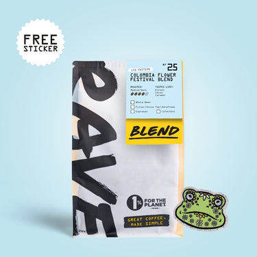 A bag of coffee with a large black-and-white graphic design, a light blue label reading Colombia Flower Festival Blend, a yellow 'BLEND' band, and a green frog-shaped sticker on a light blue background. A white burst in the top left says FREE STICKER.