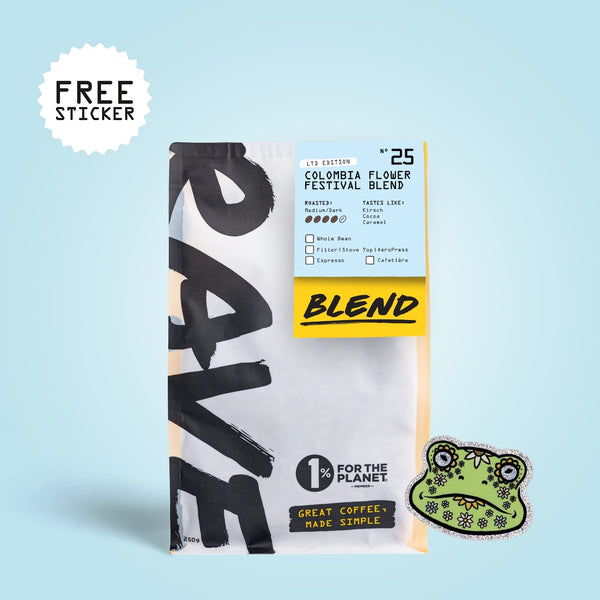 A bag of coffee with a large black-and-white graphic design, a light blue label reading Colombia Flower Festival Blend, a yellow 'BLEND' band, and a green frog-shaped sticker on a light blue background. A white burst in the top left says FREE STICKER.