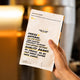 A hand holding a white coffee bag with black and yellow text that reads 'FRESH NATURAL' and 'QUALITY COFFEE' against a blurred warm-toned indoor background.