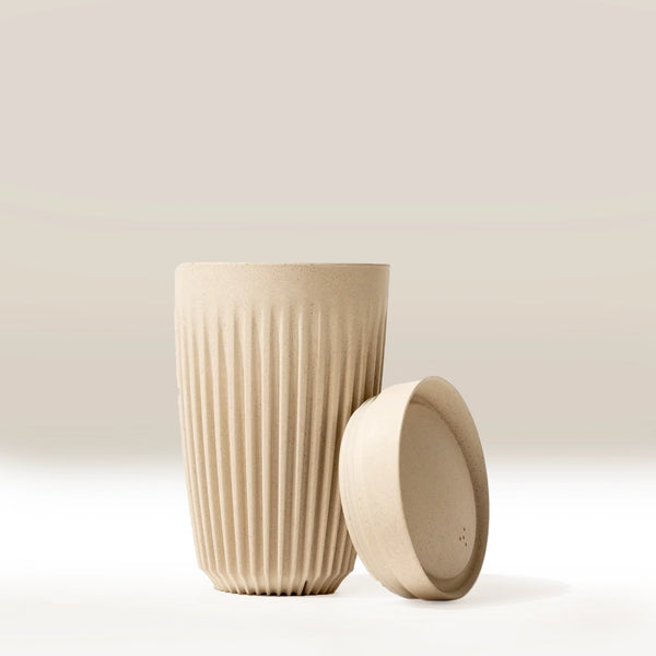 The Huskee 12oz Original coffee cup by Huskee, in beige with textured vertical grooves, stands upright next to its matching side-placed lid. A soft gradient light beige backdrop highlights this eco-friendly reusable cup.