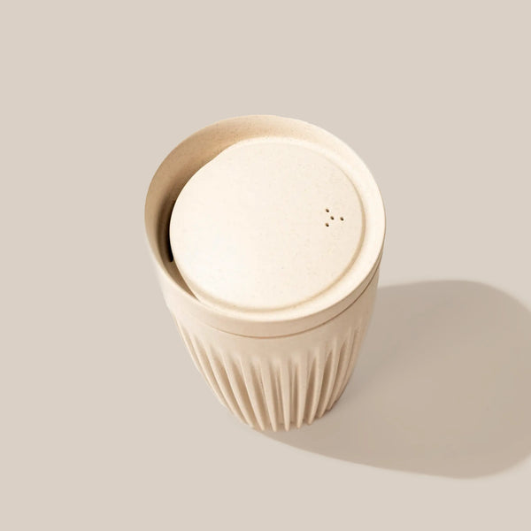 The 12oz Huskee Original by Huskee is an eco-friendly beige cup with a matching lid and vertical grooves on its lower half, displayed against a light beige background with a subtle shadow.