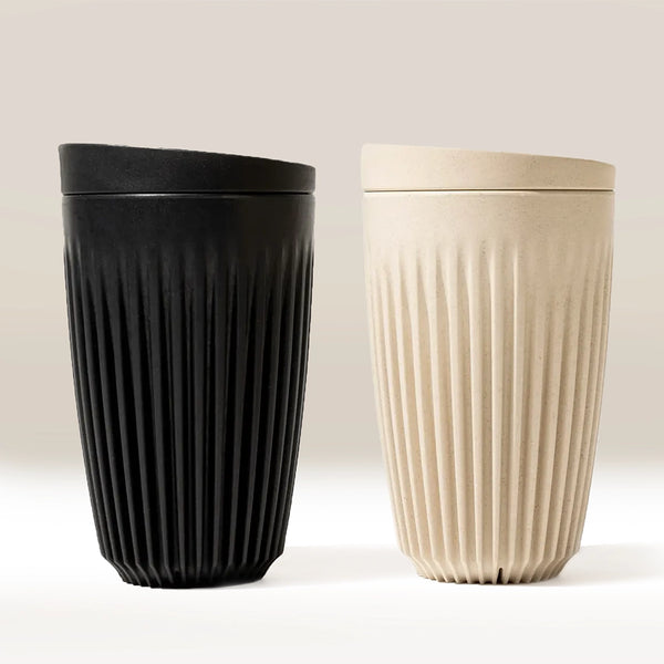 Two Huskee 12oz Original coffee cups, one black and one beige, are side by side on a neutral background. The eco-friendly Huskee cups have matching lids, vertical ridges, and a modern minimalist design.