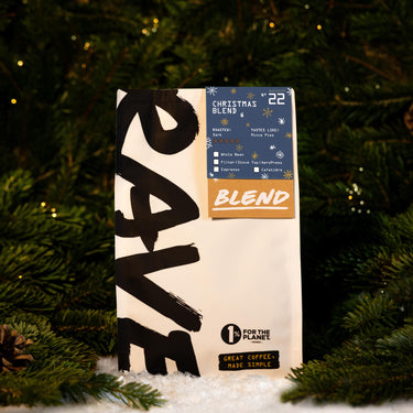A bag of RAVE COFFEE Christmas Blend Nº 22 featuring tasting notes of mince pies and a dark roast intensity, set against a background of evergreen branches.