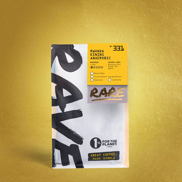 A white and yellow RAVE COFFEE bag labeled Rwanda Kinini Anaerobic Nº 331 stands on a gold background, featuring the Kinini Washing Station, fruit-forward notes, roast details, and the 1% for the Planet logo.
