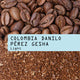 Close-up of whole coffee beans above ground coffee, separated by a blue label that reads Colombia Danilo Pérez Gesha Nº 347 by RAVE COFFEE—gesha beans from Huila, Colombia featuring distinct tropical fruit notes.