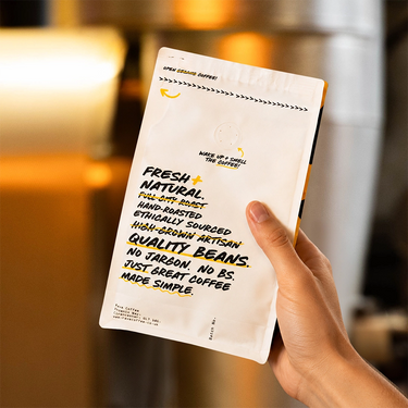 A hand holding a white coffee bag package with bold black and yellow text in a warm, blurred indoor setting.