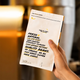A hand holds a white coffee bag package with black and yellow text in a warm, blurred indoor setting. The package reads 'FRESH NATURAL' and 'QUALITY COFFEE' with small yellow banana illustrations near the top.