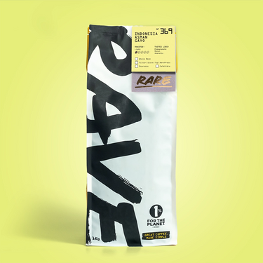 A single bag of coffee stands upright against a pale yellow-green background. The bag is mostly white with large black brushstroke lettering and a small yellow label near the top.