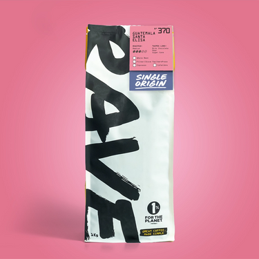 A single bag of coffee stands upright against a solid pink background. The bag is white with large black brushstroke lettering and a pink, blue, and yellow label area near the top.