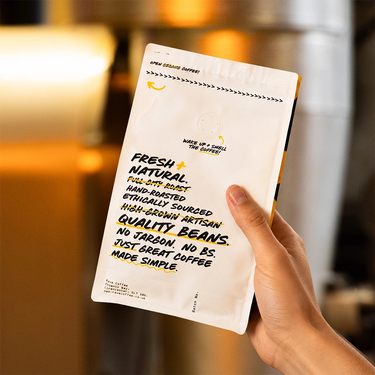 A hand holds a white coffee bag package with black and yellow text in a warm, blurred indoor setting. The package reads 'FRESH NATURAL' and 'QUALITY COFFEE' with small yellow banana illustrations near the top.