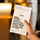 A hand holds a white coffee bag package with black and yellow text in a warm, blurred indoor setting. The package reads 'FRESH NATURAL' and 'QUALITY COFFEE' with small yellow banana illustrations near the top.