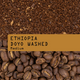 Coffee bean product graphic showing roasted coffee beans at the bottom, coarse ground coffee at the top, and a tan label across the center reading “ETHIOPIA DOYO WASHED Medium.”.