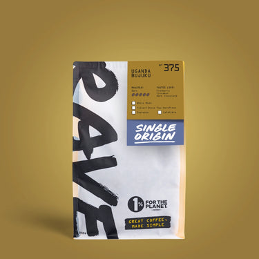 A bag of One Coffee Luganda Buuku single origin coffee stands upright against a solid mustard-yellow background. The packaging is white with bold black brushstroke lettering on the left, a gold label at the top right, and a blue banner reading 'Single Origin'.