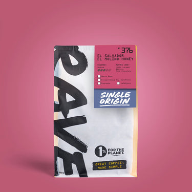 A bag of El Salvador El Rolino Honey coffee on a solid pink background. The packaging features a white and black brush-stroke design with a pink and blue label section detailing the single-origin coffee from Maní Coffee Roasters.