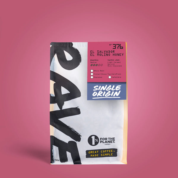 A bag of El Salvador El Rolino Honey coffee on a solid pink background. The packaging features a white and black brush-stroke design with a pink and blue label section detailing the single-origin coffee from Maní Coffee Roasters.
