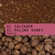 Coffee beans and ground coffee arranged with a pink label across the middle reading 'EL SALVADOR EL MOLINO HONEY Medium'.