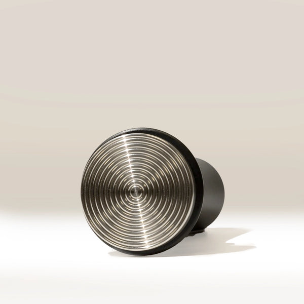 A matte black cylindrical Self Levelling Spring Tamper by RAVE Coffee, featuring a metallic concentric circular pattern on its front, is shown lying on its side against a plain light background, sized for a 54mm espresso filter basket.