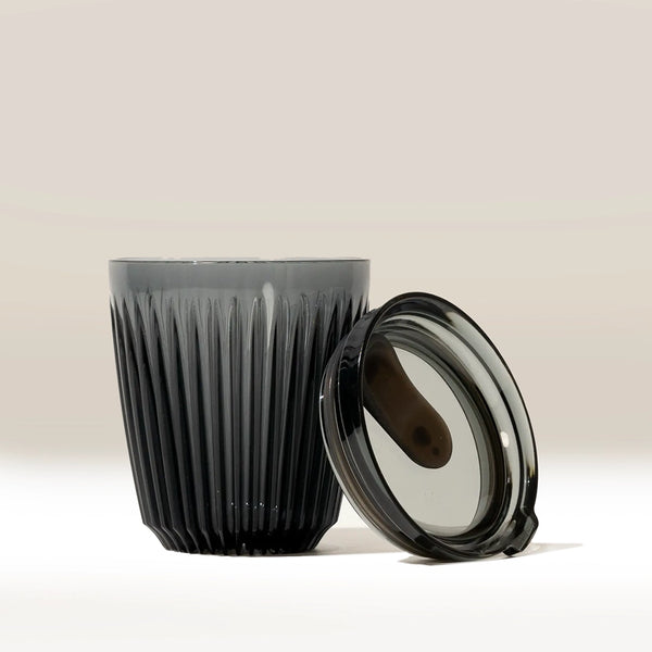 The Huskee 8oz Huskee Renew, a dark gray ribbed reusable coffee cup by Huskee, made from Tritan Renew 50, is displayed with a matching transparent lid leaning against it on a neutral background.