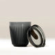 The Huskee 8oz Huskee Renew, a dark gray ribbed reusable coffee cup by Huskee, made from Tritan Renew 50, is displayed with a matching transparent lid leaning against it on a neutral background.