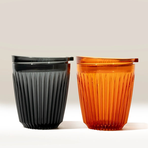 Two ribbed, translucent 8oz Huskee Renew reusable coffee cups by Huskee are shown side by side, one in dark gray and one in orange, each with a matching lid. Both rest on a light surface against a neutral background.