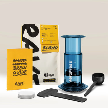 Aeropress Gift Set Clear/Colour by Aeropress: This comprehensive home coffee brewing set features the Aeropress coffee maker with plunger, scoop, stirrer, filter papers, a yellow brew guide, and a bag of Rave Signature Blend coffee—artfully presented on a light background. An ideal gift for any coffee enthusiast.