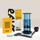 Aeropress Gift Set Clear/Colour by Aeropress: This comprehensive home coffee brewing set features the Aeropress coffee maker with plunger, scoop, stirrer, filter papers, a yellow brew guide, and a bag of Rave Signature Blend coffee—artfully presented on a light background. An ideal gift for any coffee enthusiast.