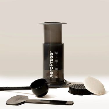 The Aeropress AeroPress Coffee Maker by Aeropress comes with a scoop, stirrer, filter holder, and round paper filters—neatly arranged on a light surface with a neutral background—ideal for brewing coffee at home.