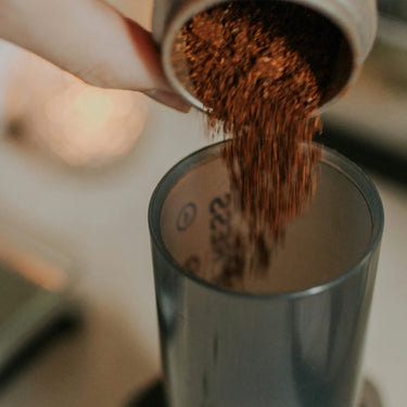 A hand pours ground coffee from a container into an Aeropress AeroPress Coffee Maker, ready for home brewing, set against a softly blurred background.