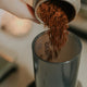 A hand pours ground coffee from a container into an Aeropress AeroPress Coffee Maker, ready for home brewing, set against a softly blurred background.