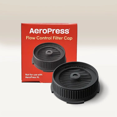 The black AeroPress Flow Control Filter Cap by Aeropress is displayed before its red box, which features white text and the cap image. The box notes it’s not for AeroPress XL. The pressure-actuated valve enables espresso-like crema.