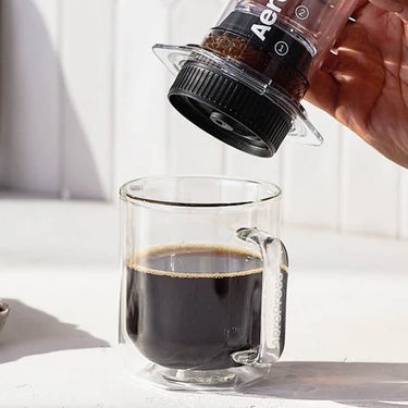 A hand presses an AeroPress coffee maker with the AeroPress Flow Control Filter Cap by Aeropress onto a glass mug of black coffee, sunlight streaming in as rich crema is about to form.