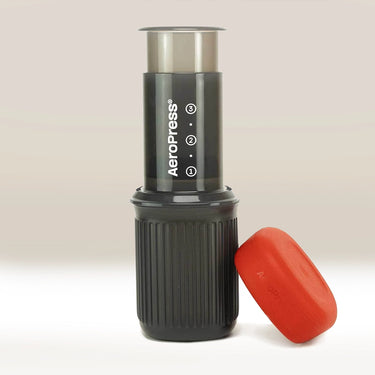 The Aeropress AeroPress Go travel coffee maker, featuring a black and gray design with a red cap resting beside its base, stands upright on a light surface. The compact brewer displays the AeroPress logo and instructions.