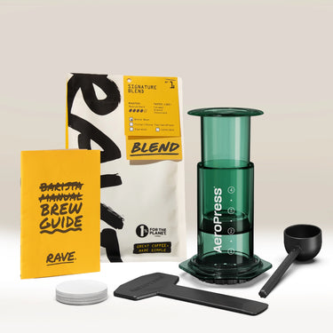 Introducing the Aeropress Gift Set Clear/Colour by Aeropress—a thoughtfully curated gift for coffee lovers. This set includes a home coffee brewer with an AeroPress, a premium blend bag of coffee, brew guide booklet, scoop, stirrer, filter papers, and filter holder—all beautifully arranged on a white surface. The perfect present for any coffee enthusiast.