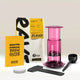 An Aeropress Gift Set Clear/Colour by Aeropress—including a vibrant pink coffee maker, black scoop, stirrer, paper filters, a cheerful yellow brew guide, and a bag of Rave coffee beans—is beautifully arranged on a neutral background, making it the perfect gift for any coffee enthusiast or home brewing aficionado.