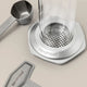 A close-up of the Aerobie AeroPress Premium Coffee Maker with metal filter, metal scoop, and stirrer on a light surface. Both the coffee maker and scoop feature Aerobie branding, highlighting its 3-in-1 brewing technology.
