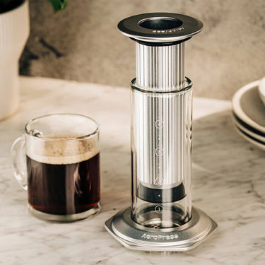 A clear glass mug filled with black coffee sits beside the Aerobie AeroPress Premium Coffee Maker on a marble countertop, with plates and a plant in the background.