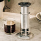 A clear glass mug filled with black coffee sits beside the Aerobie AeroPress Premium Coffee Maker on a marble countertop, with plates and a plant in the background.