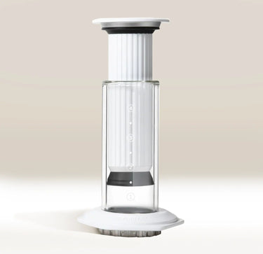 A clear and white AeroPress Premium Coffee Maker by Aerobie stands upright on a light surface, highlighting its plunger, filter cap, measurement markings, and innovative 3-in-1 brewing technology.