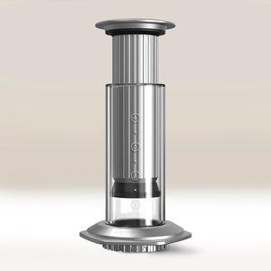 The Aerobie AeroPress Premium Coffee Maker, featuring a silver and clear cylindrical design with measurement markings and 3-in-1 brew technology, stands upright on a light surface against a neutral background.