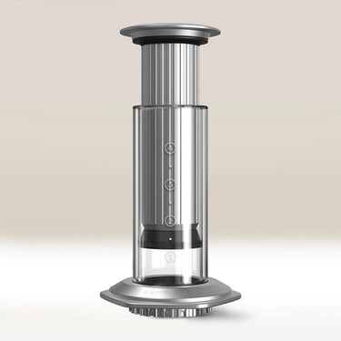 The Aerobie AeroPress Premium Coffee Maker, featuring a silver and clear cylindrical design with measurement markings and 3-in-1 brew technology, stands upright on a light surface against a neutral background.