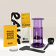 An AeroPress Gift Set Clear/Colour by AeroPress, featuring a vibrant purple AeroPress, coffee scoop, stirrer, paper filters, a bag of Rave Signature Blend coffee, and a bright yellow brew guide, is neatly arranged on a plain surface against a light background—everything you need for the perfect home coffee brewing experience. This set makes an ideal gift for both coffee enthusiasts and beginners.